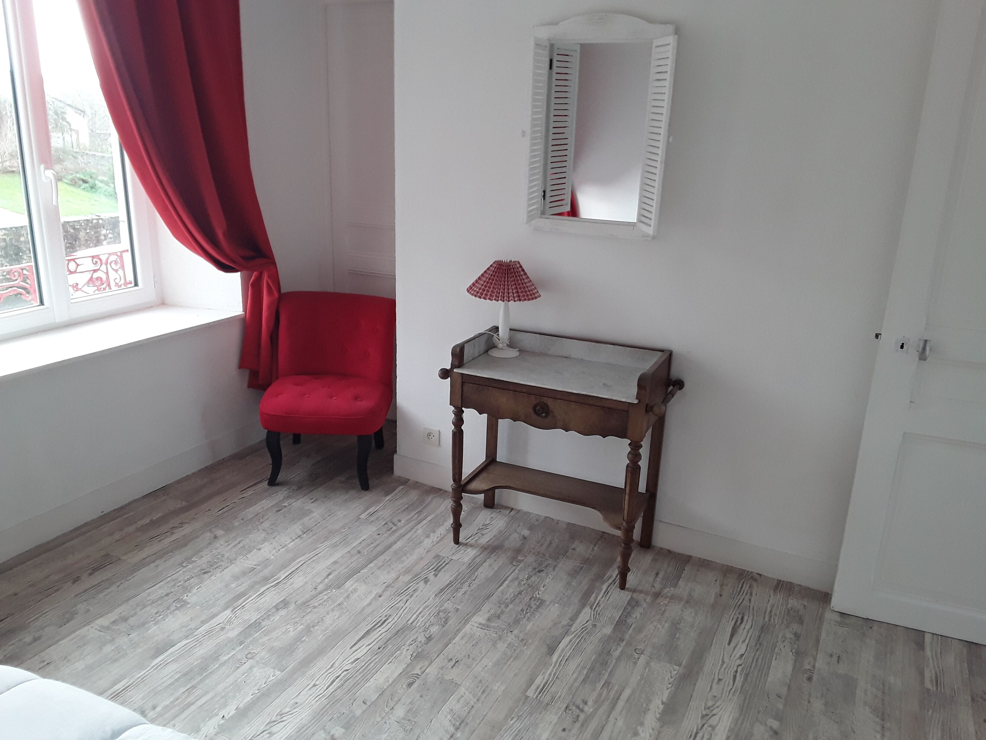 A room with a wooden floor, a red chair, a small wooden table with a lamp, and a window with red curtains.