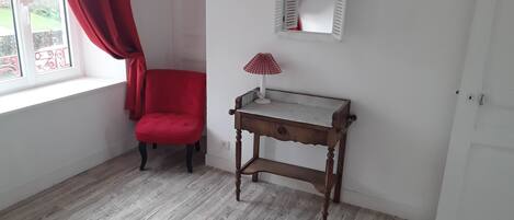 A room with a wooden floor, a red chair, a small wooden table with a lamp, and a window with red curtains.