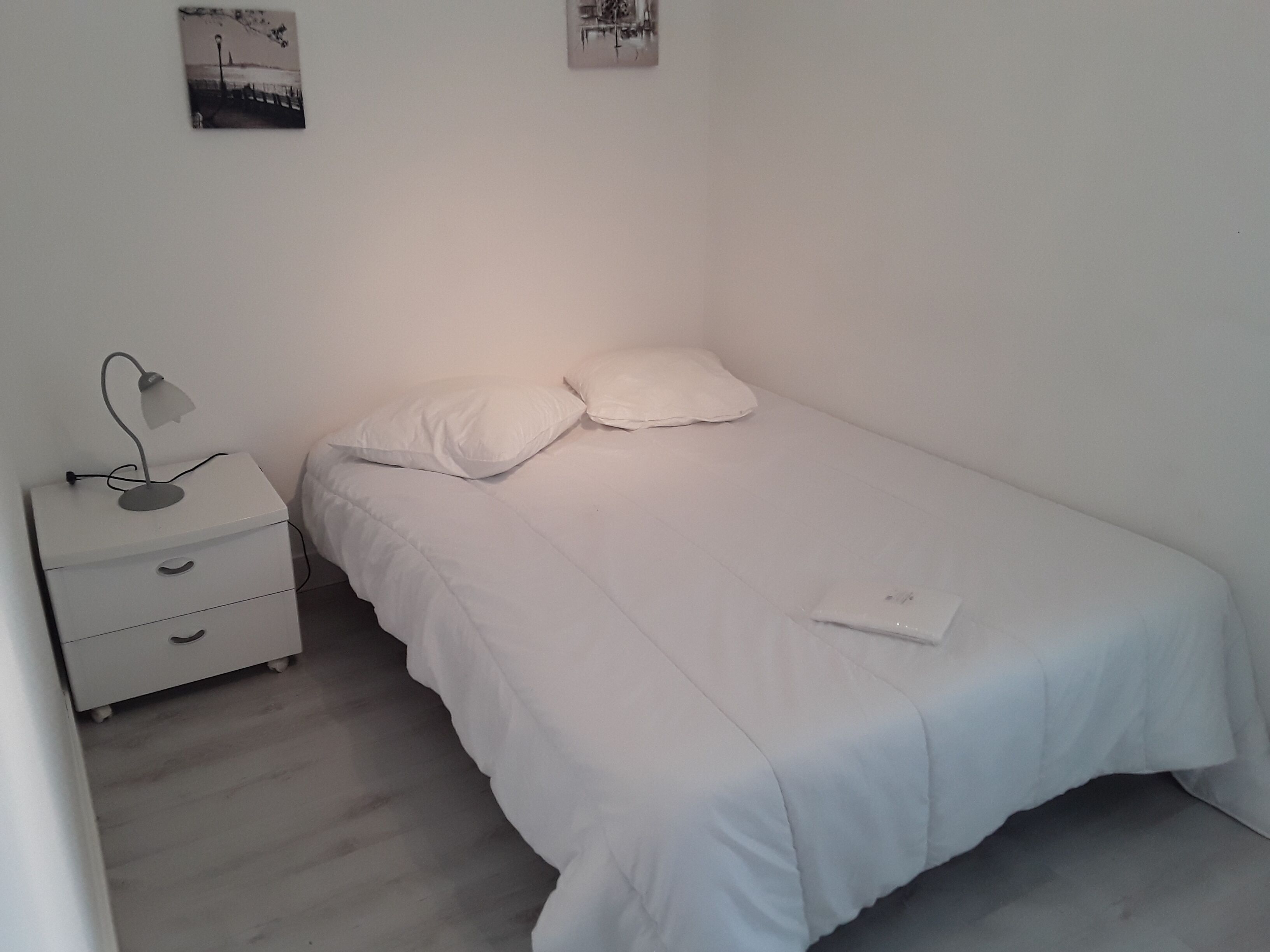 Quarto pequeno e branco com uma cama de solteiro, mesa de cabeceira, abajur e duas almofadas.