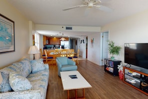 A living room with a sofa, a coffee table, a TV, and a bookshelf. There is a dining area with a table and chairs.