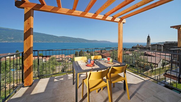 Una terraza al aire libre con una pérgola de madera, una mesa dispuesta para comer y vistas al mar y edificios a lo lejos.
