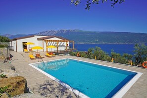 A swimming pool with lounge chairs and umbrellas, a building with a view of the sea, and mountains in the background. - Residence Dany (Gargnano)