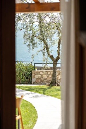 A view through a doorway to a patio area with a tree, a stone wall, and a pathway leading to a body of water. - Residence Dany (Gargnano)