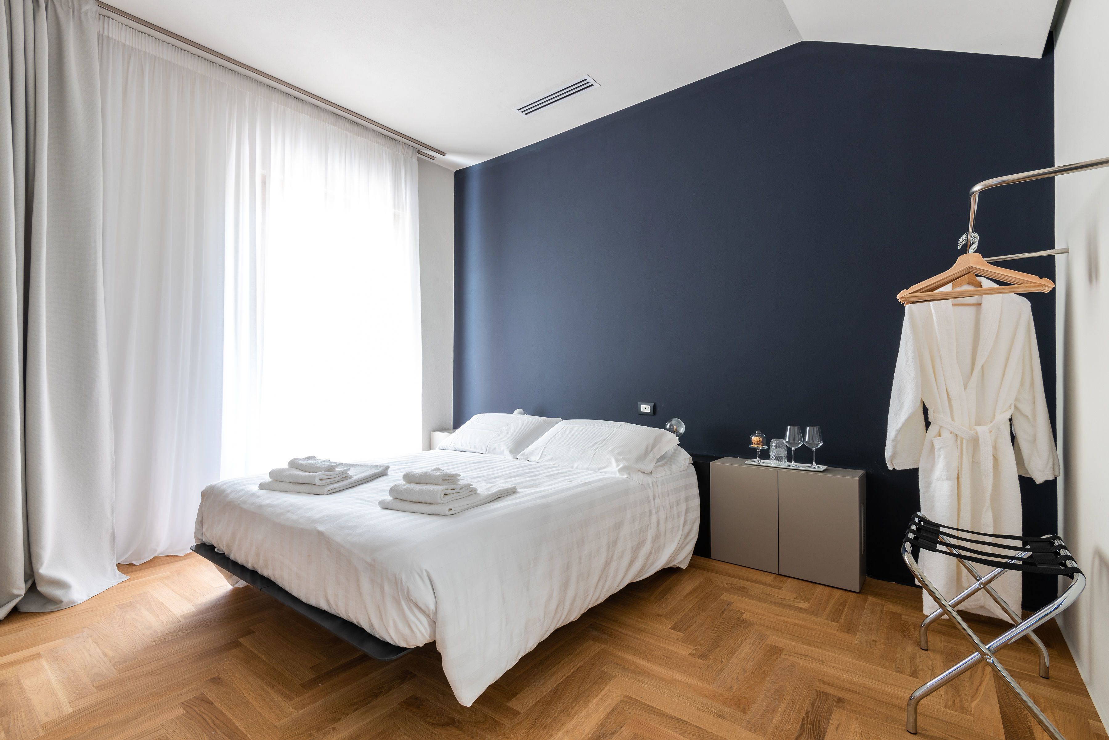 A modern hotel room with a large bed, a bedside table with a lamp, a small nightstand, a wooden hanger, and a white robe on a hook.