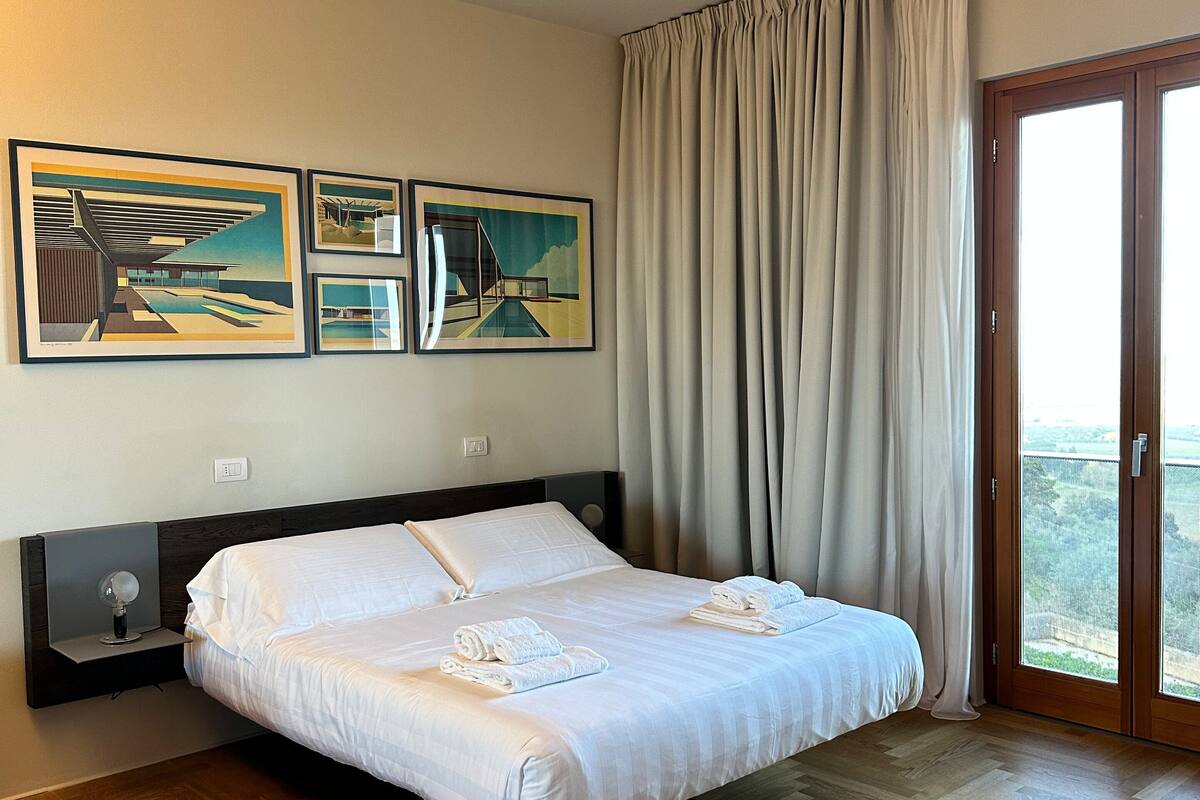 A hotel room with a large bed, a wooden floor, a balcony with curtains, and three framed artworks on the wall.