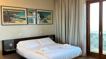 A hotel room with a large bed, a wooden floor, a balcony with curtains, and three framed artworks on the wall.