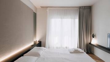 Una habitación de hotel moderna con una cama grande, una mesita de noche, un televisor y un ventanal con cortinas de gasa.