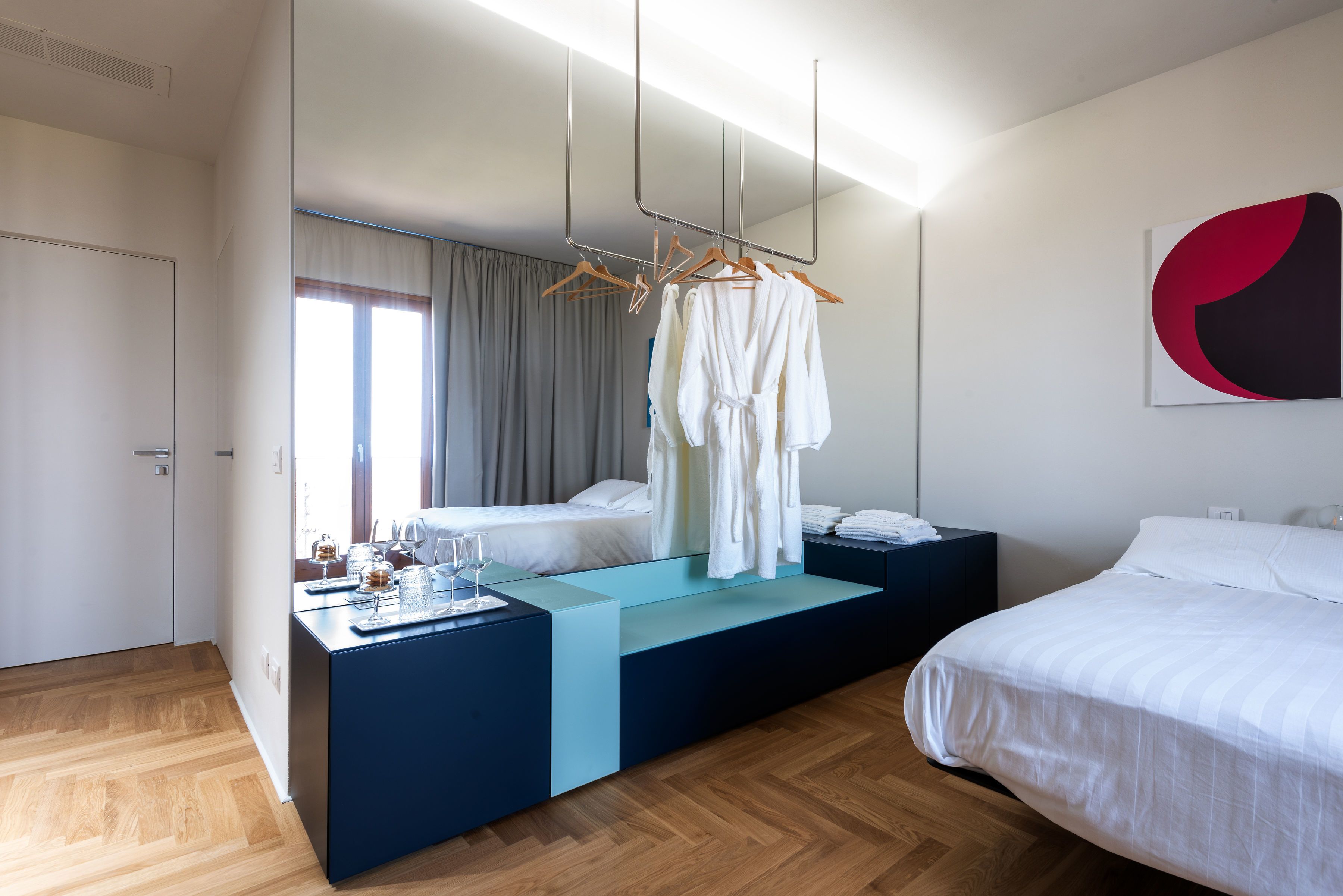 A modern hotel room with a wooden floor, a bed with white bedding, a bedside table with a lamp, and a large window with curtains.