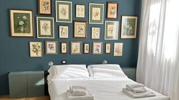 Una cama bien tendida con ropa de cama y toallas blancas, una pared azul con impresiones botánicas enmarcadas y un suelo de madera en patrón espiga.