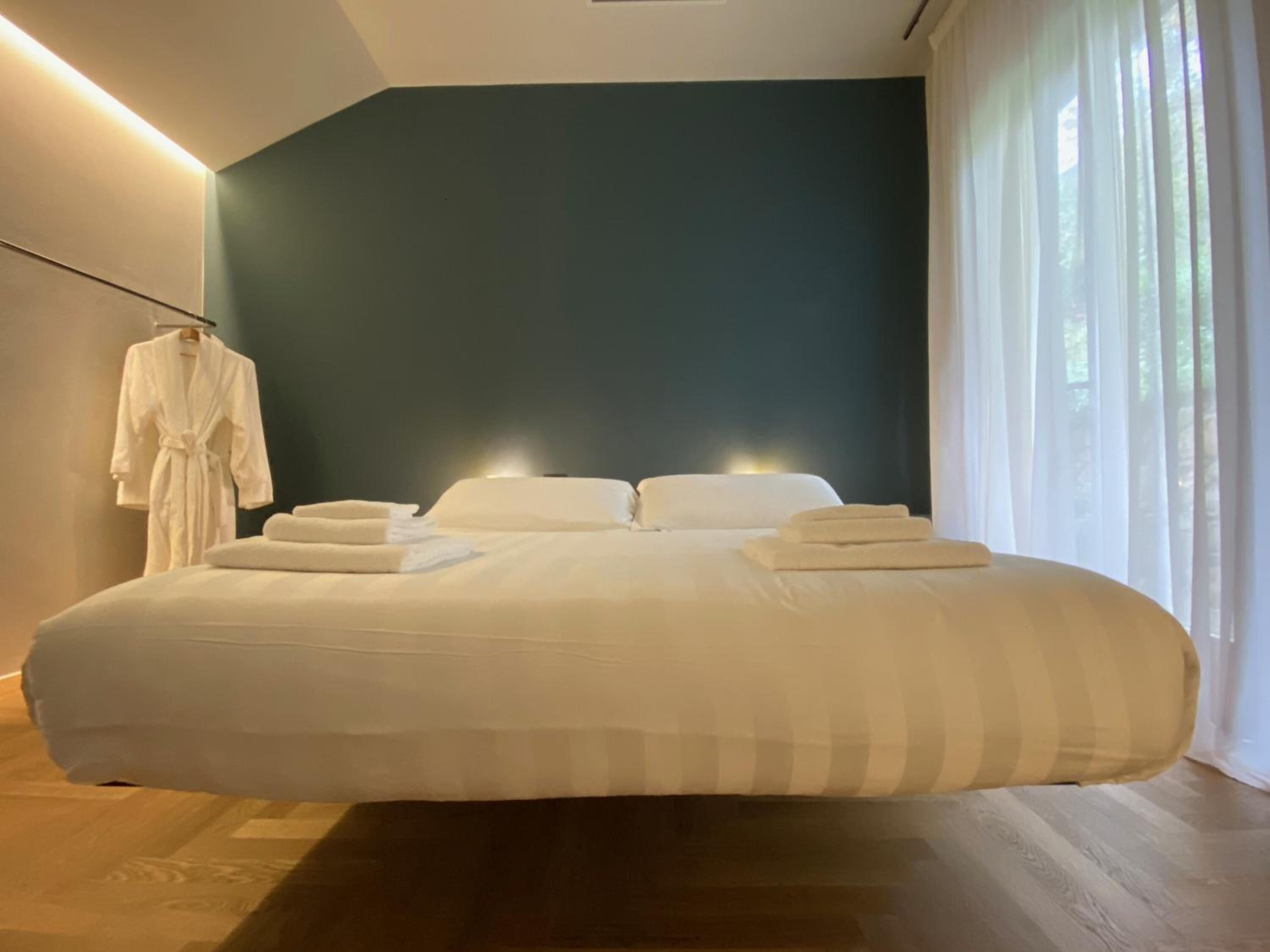 A modern bedroom with a large bed, a robe hanging on a rack, and a view of greenery through sheer curtains.