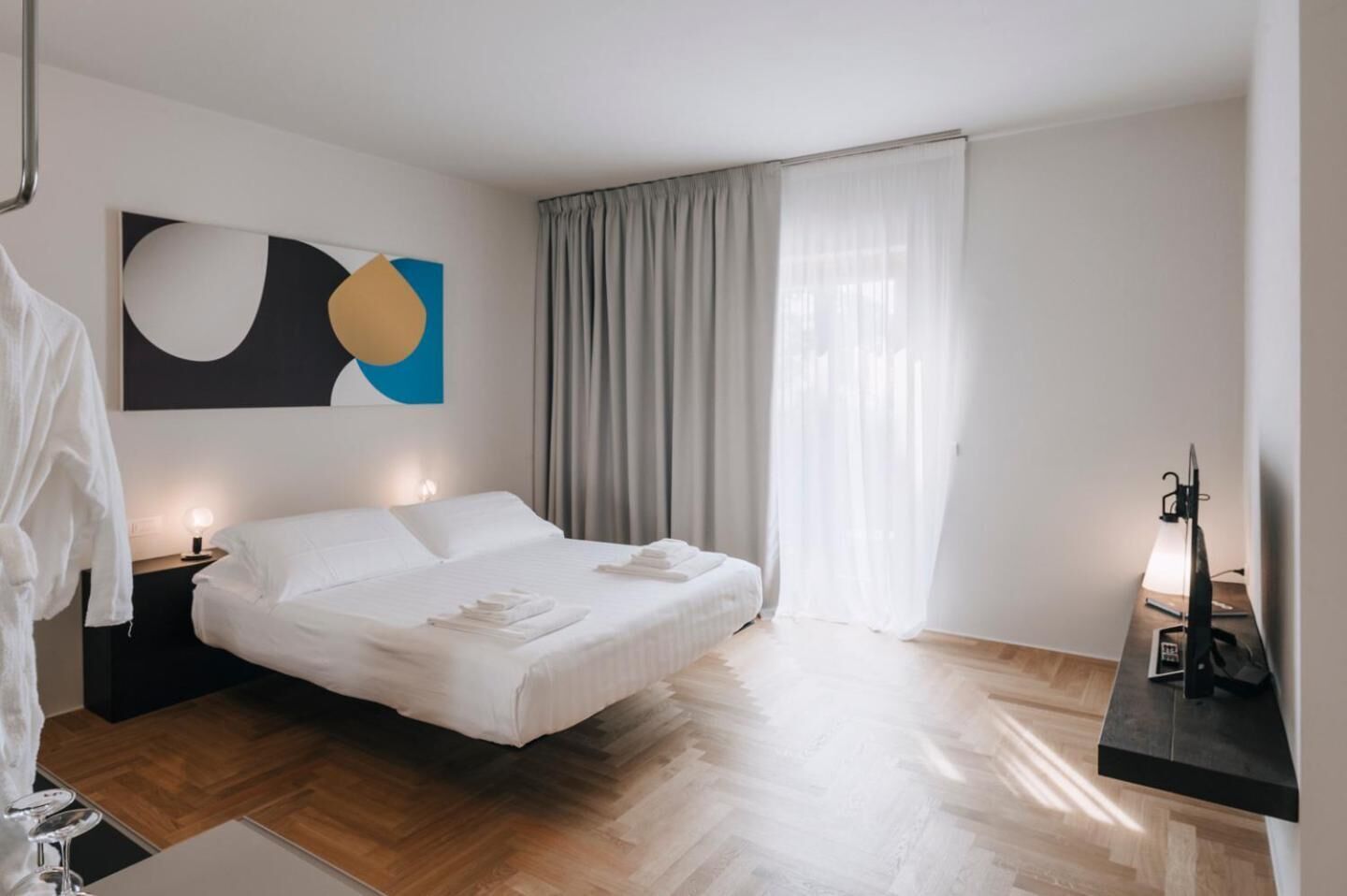 Ein modernes Hotelzimmer mit einem großen Bett, einem Nachttisch mit Lampe, einem Schreibtisch mit Computer und einem großen, abstrakten Gemälde an der Wand.