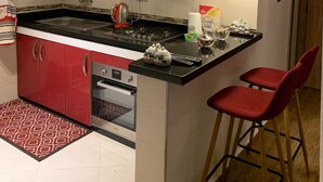 A compact kitchen with red cabinets, a black countertop, a gas stove, and a small island with two red bar stools. - Appartement Lalla Ghita (Essaouira)