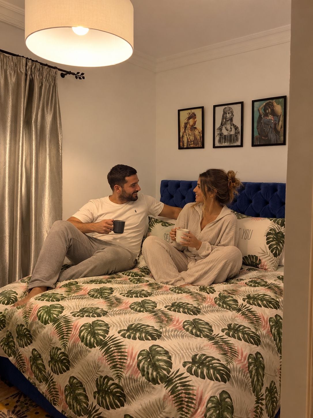Una pareja sentada en una cama con un edredón estampado con hojas tropicales, un cabecero azul y tres cuadros en la pared.