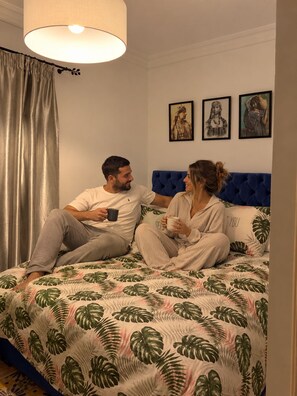 A couple sitting on a bed with a tropical leaf pattern bedspread, a blue headboard, and three framed artworks on the wall. - Appartement Lalla Ghita (Essaouira)