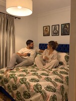 Una pareja sentada en una cama con un edredón estampado con hojas tropicales, un cabecero azul y tres cuadros en la pared.