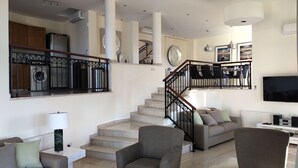 A spacious living area with a staircase, a sofa, a coffee table, and a TV. - Villa HERMIONA - Wake Up To A Golf View (Paphos)
