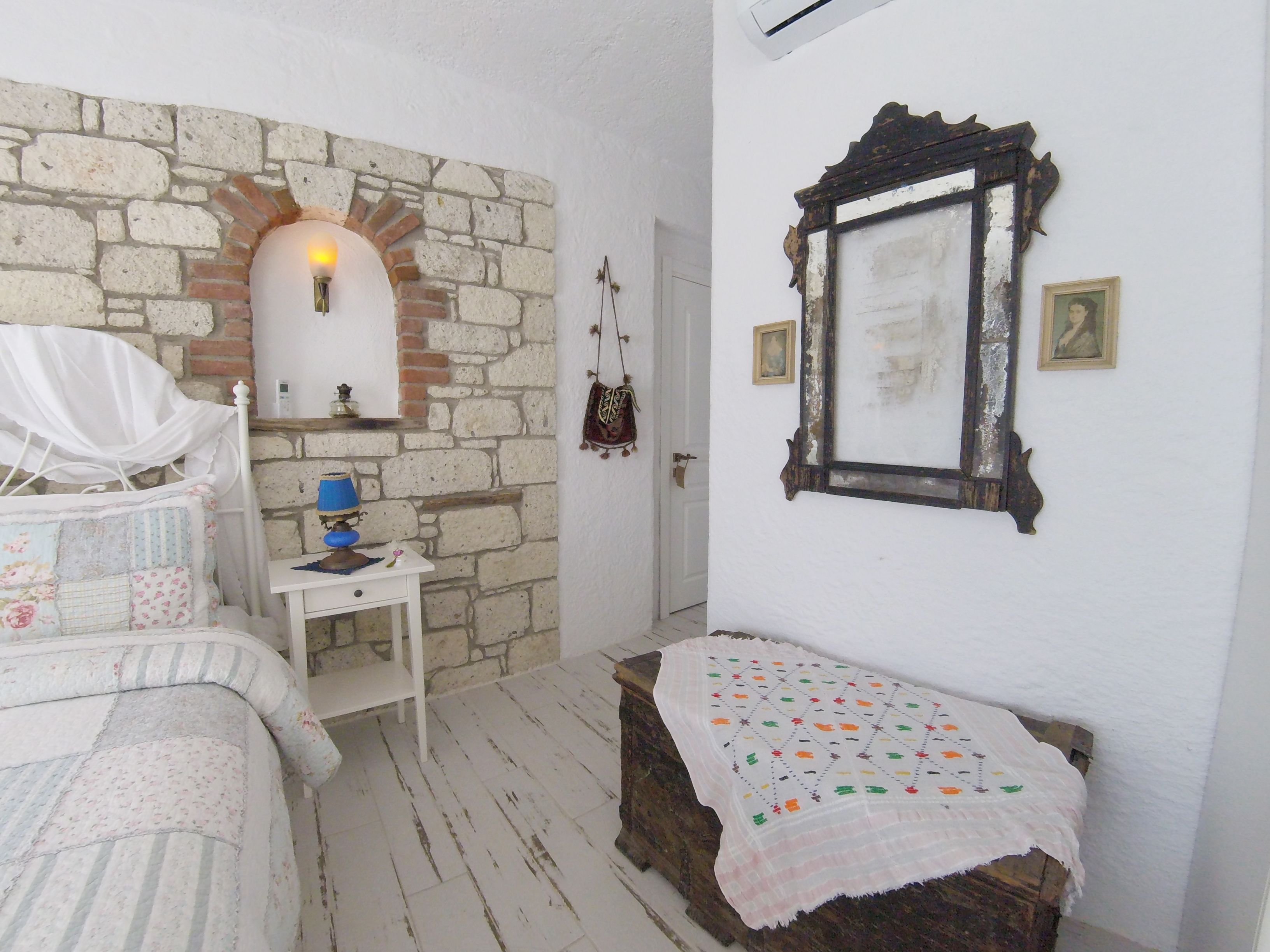 A bedroom with a stone wall, a bed with a floral headboard, a bedside table with a lamp, and a mirror on the wall.
