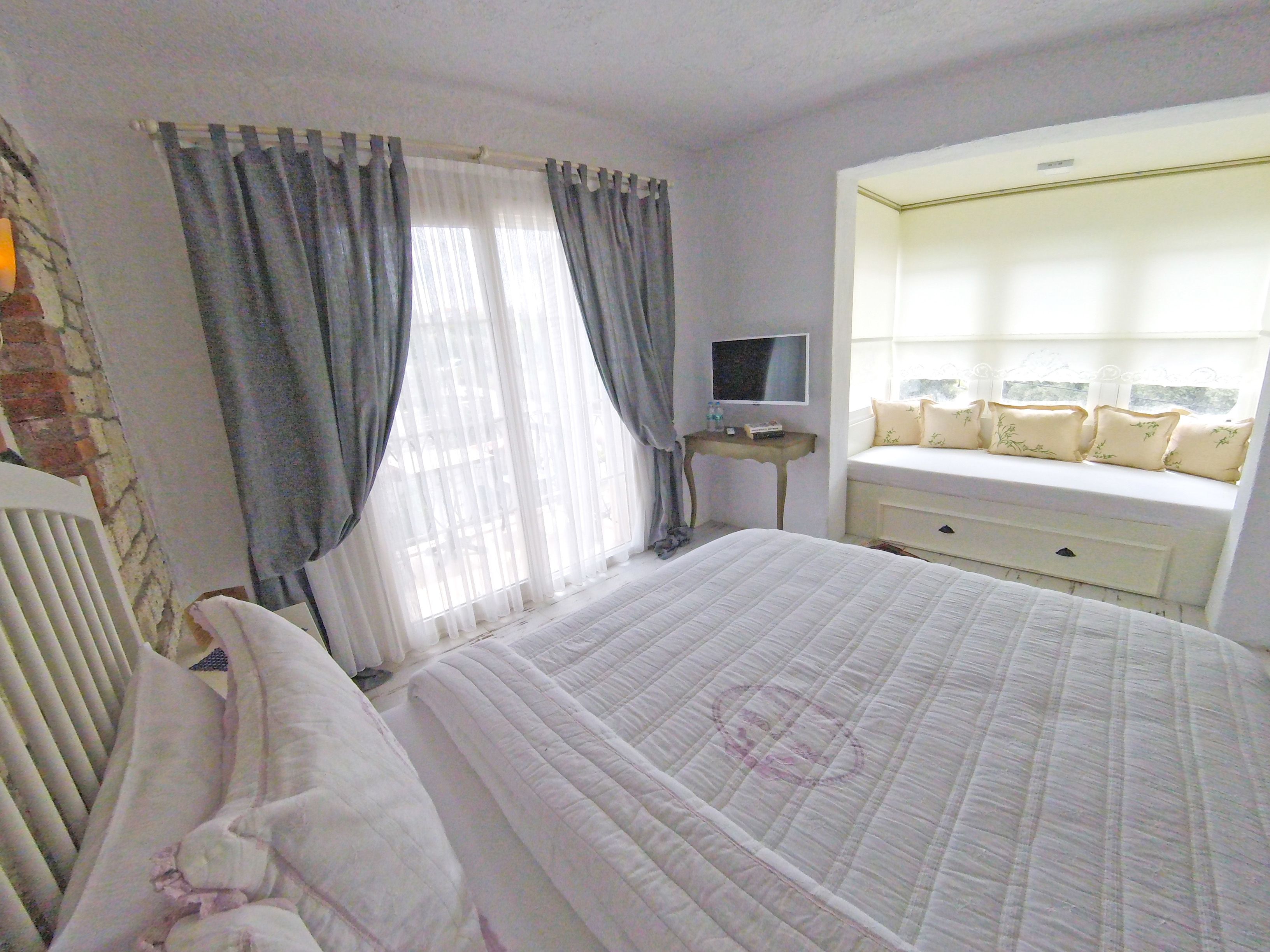Quarto com cama grande, mesa de cabeceira com TV, janela com cortinas e um banco embutido com almofadas.