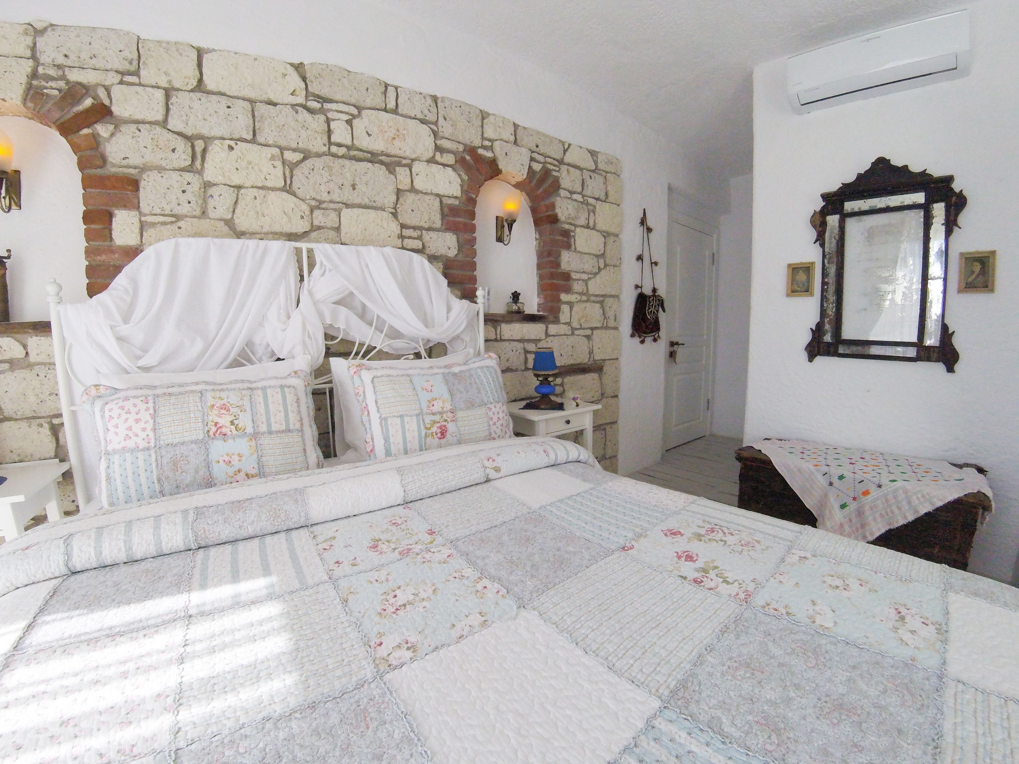 Un dormitorio con pared de piedra, cama con ropa de cama floral, mesita de noche con lámpara y espejo en la pared.