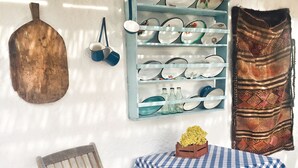 Ein rustikaler Innenraum mit einem Holzstuhl, einem Tisch mit kariertem Tischläufer, einem blauen Regal mit Geschirr und einem hängenden Topf.