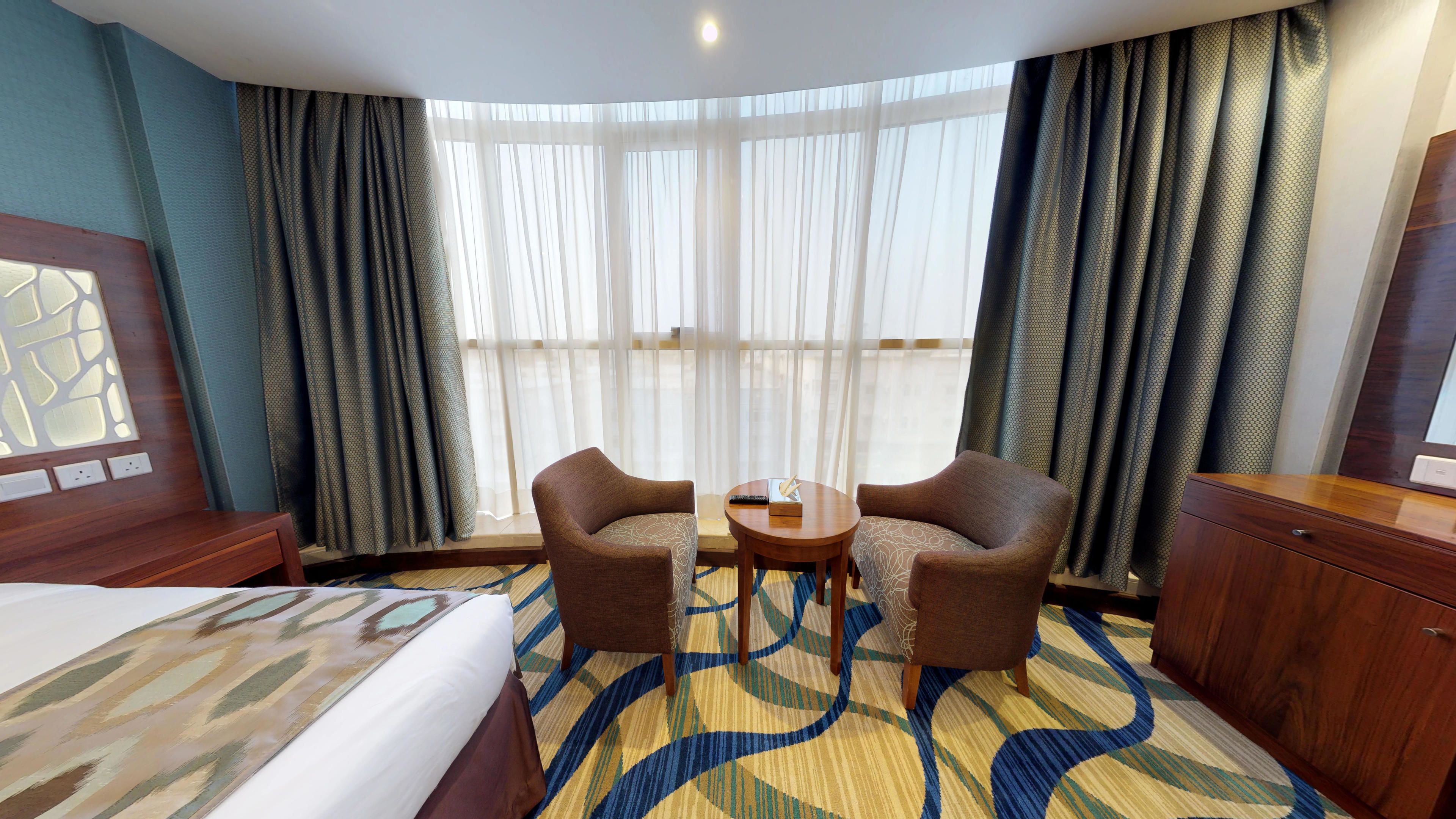 Een hotelkamer met een bed, twee fauteuils, een bijzettafeltje, een houten dressoir en een groot raam met dunne gordijnen.