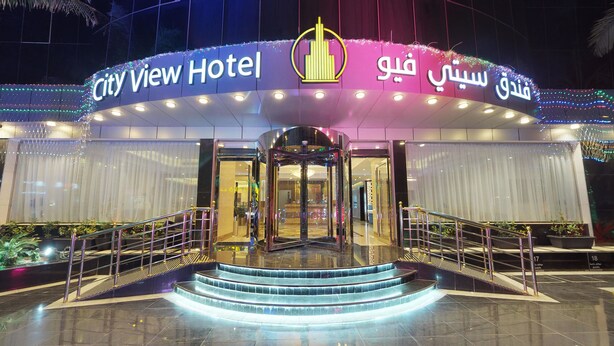 L’entrée de l’hôtel City View, avec sa porte tournante, ses marches illuminées et un panneau indiquant le nom de l’hôtel dans plusieurs langues.