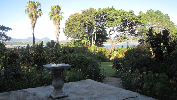 Un jardín con un pedestal de piedra, abundante vegetación y vistas a una playa y montañas al fondo.