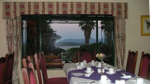 Un comedor con una mesa dispuesta para cuatro, vistas a montañas y una masa de agua, y una planta tropical en un jarrón.
