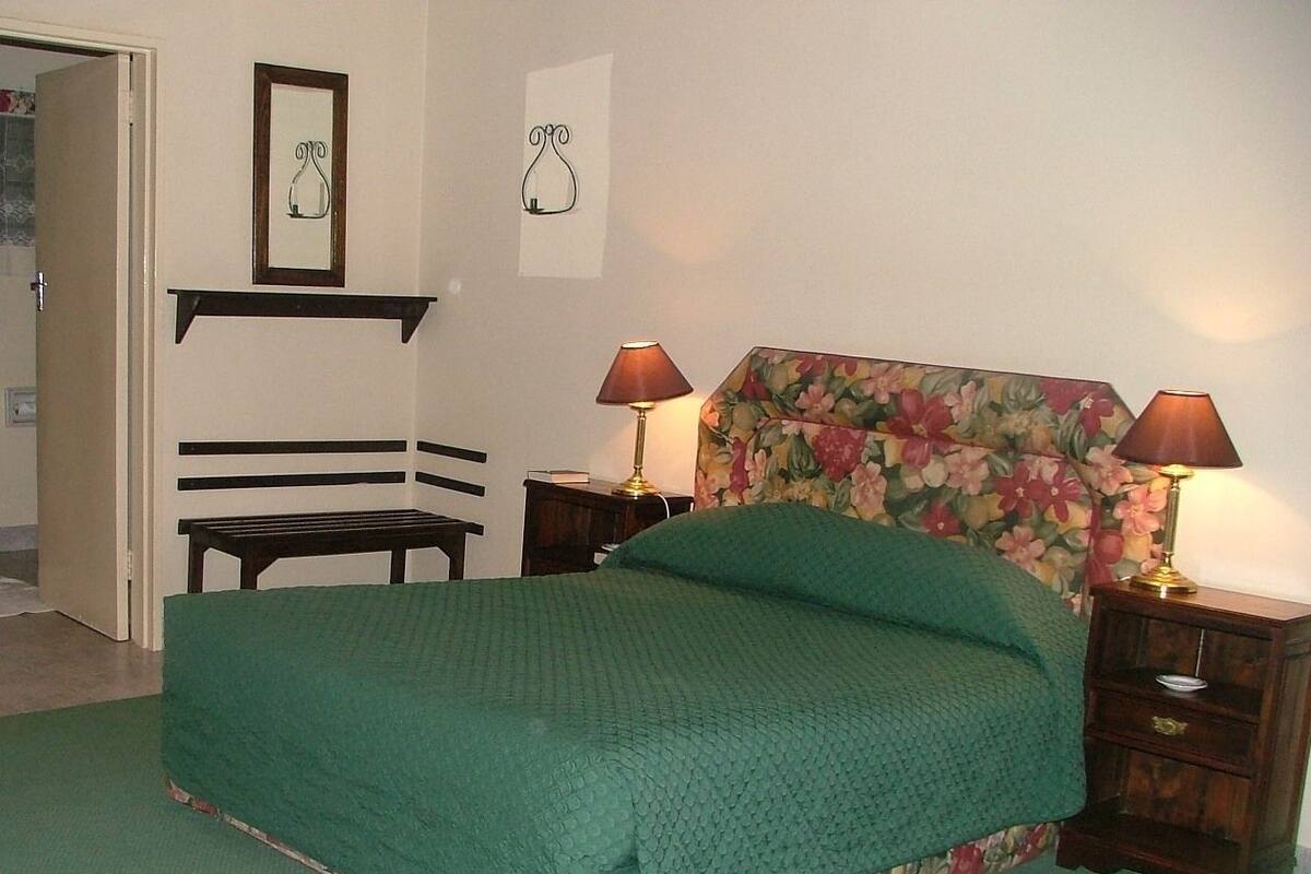 A bedroom with a green bedspread, two bedside tables with lamps, a floral headboard, a wooden bench, and a door leading to another room.