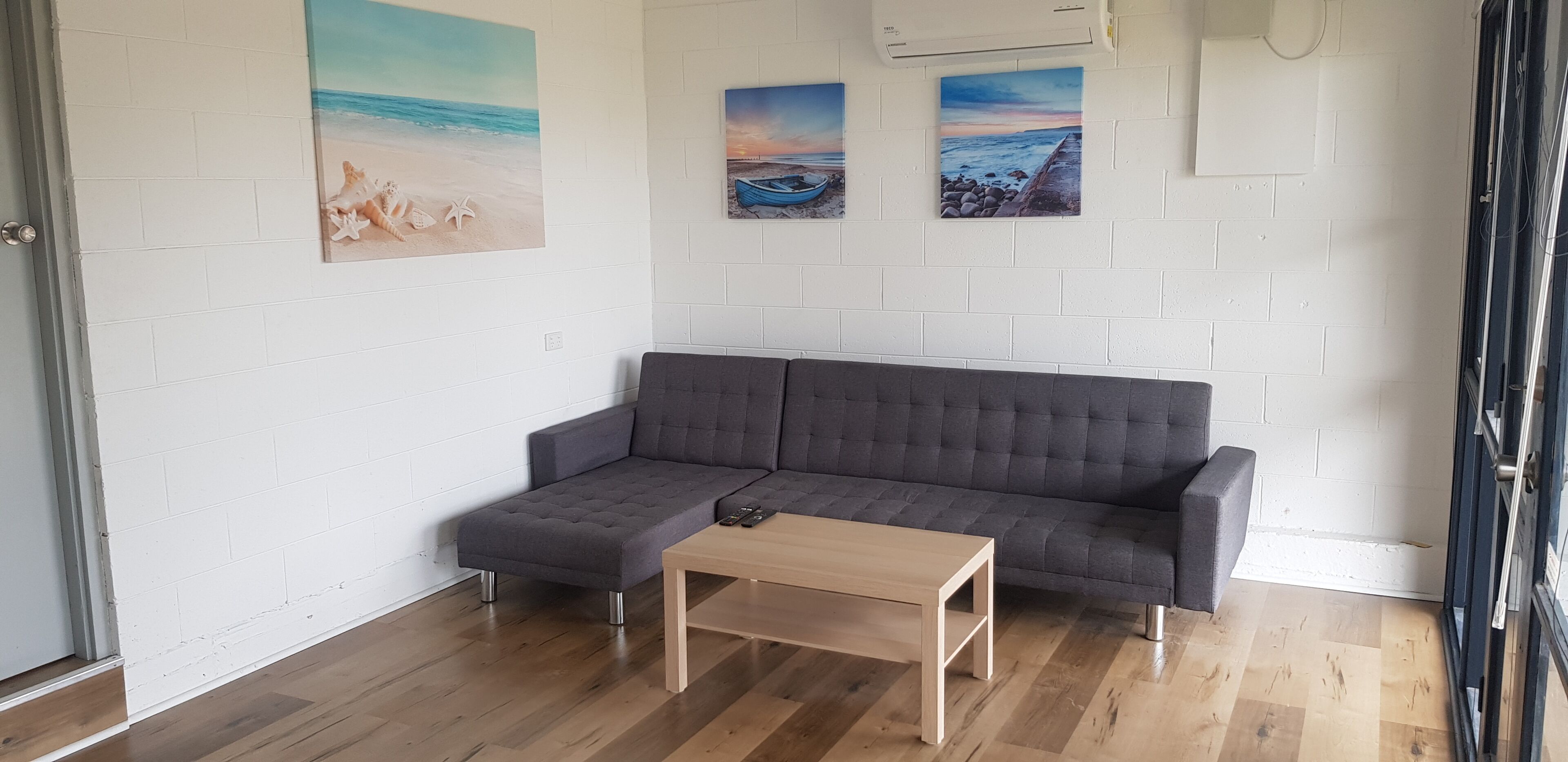 A modern living room with a grey sofa, a wooden coffee table, and three beach-themed paintings on the wall.