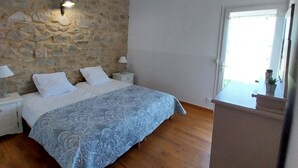 Une chambre avec un mur en pierre, un lit recouvert de draps blancs, une table de chevet avec une lampe et une fenêtre offrant une vue.