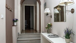 Een hal met een witte vloer, een witte muur, een bruine deur, een witte console tafel met een spiegel, een potplant en een trap naar een andere kamer.