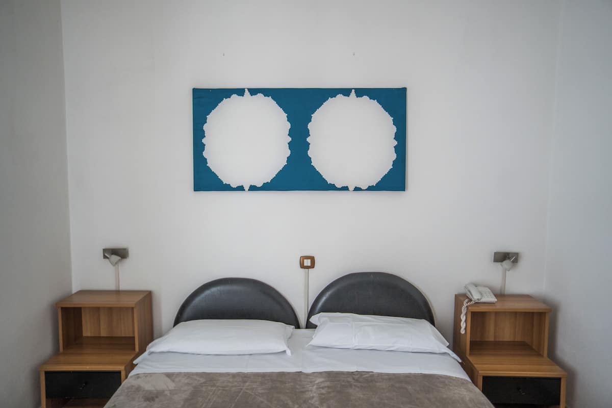 A bedroom with a bed, two bedside tables, a wall-mounted light, and a blue artwork.