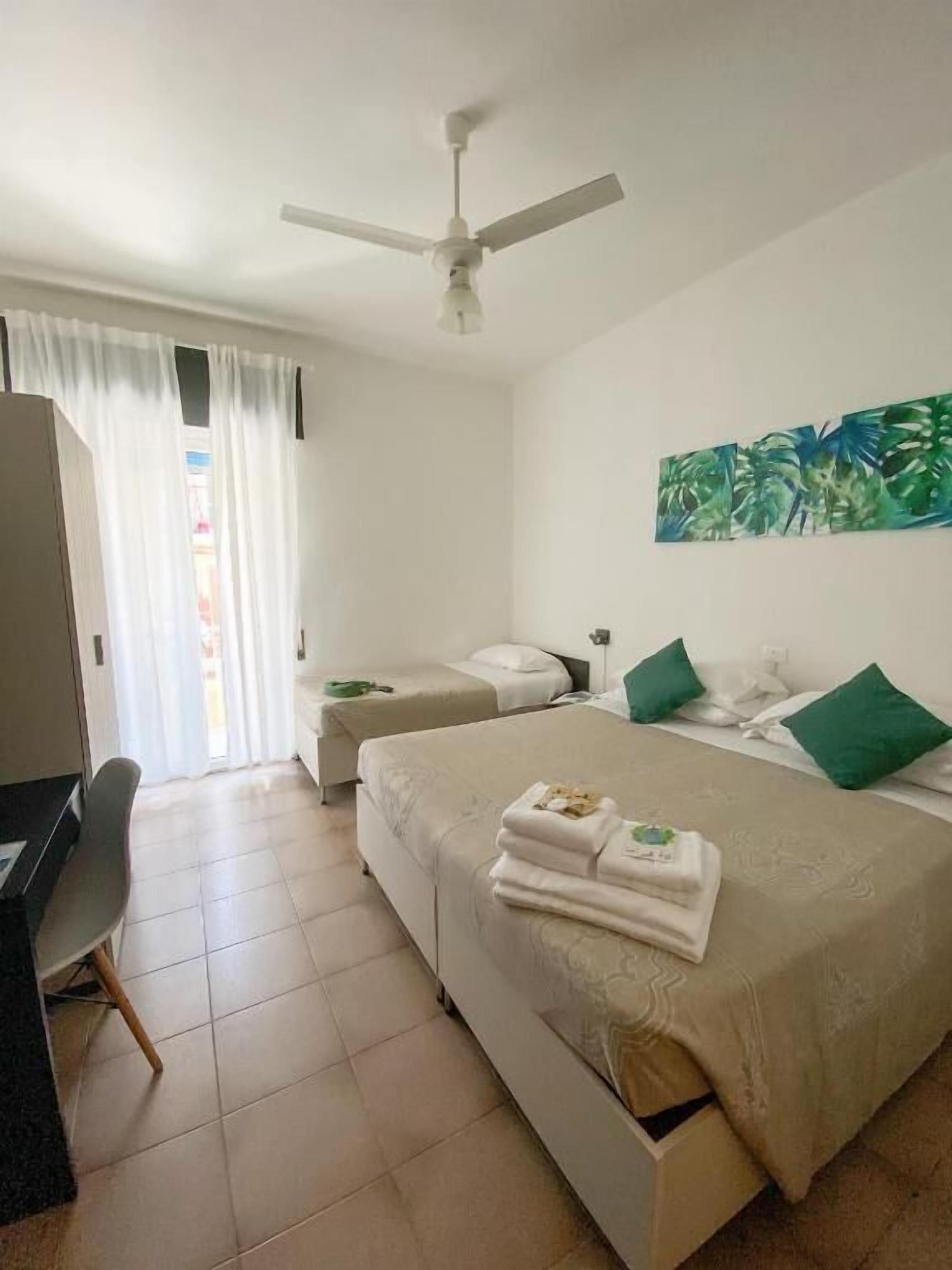A hotel room with a bed, a desk, a ceiling fan, and a painting on the wall.