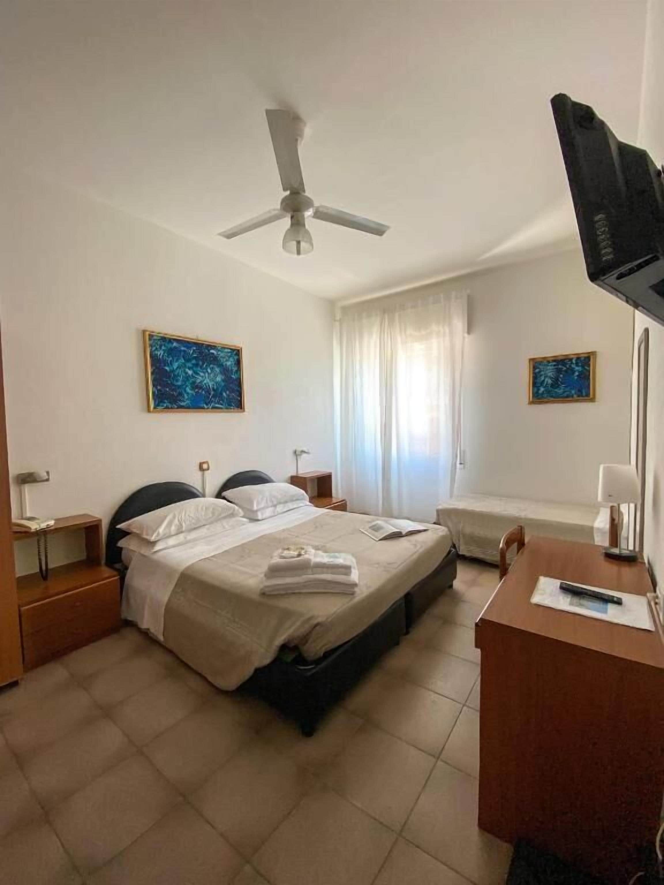 Una camera d'albergo con un letto, comodini, una scrivania, una ventola a soffitto e opere d'arte alle pareti.