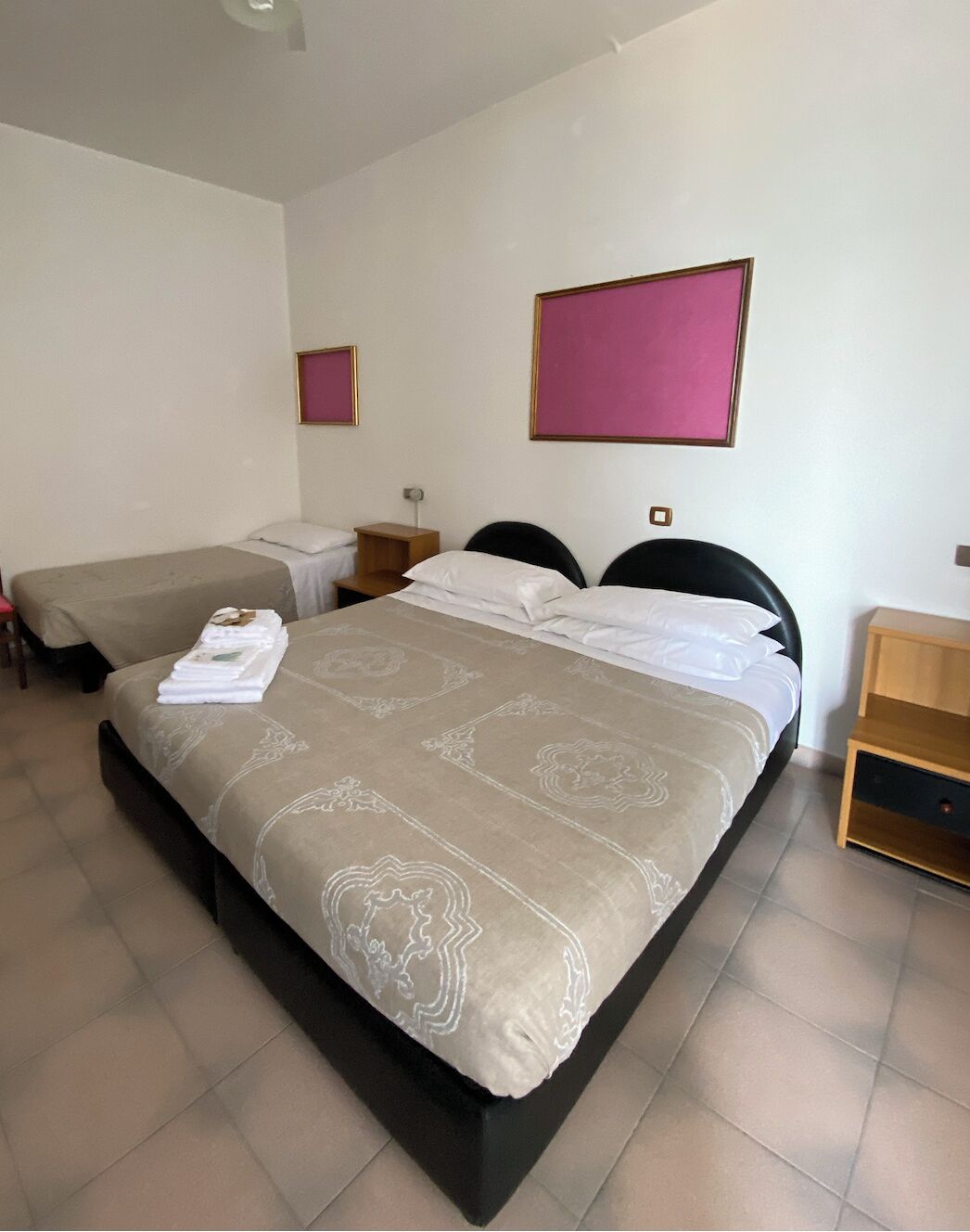 A hotel room with a large bed, two smaller beds, a wooden nightstand, and a pink wall decoration.