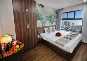 A hotel room with a bed, a nightstand, a lamp, a fruit bowl, a city view through the window, and a wall with a tropical leaf mural. - Mega Light Hotel (Nha Trang)