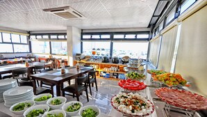 A buffet setup with various food items and a city view through large windows. - Mega Light Hotel (Bac Nha Trang)