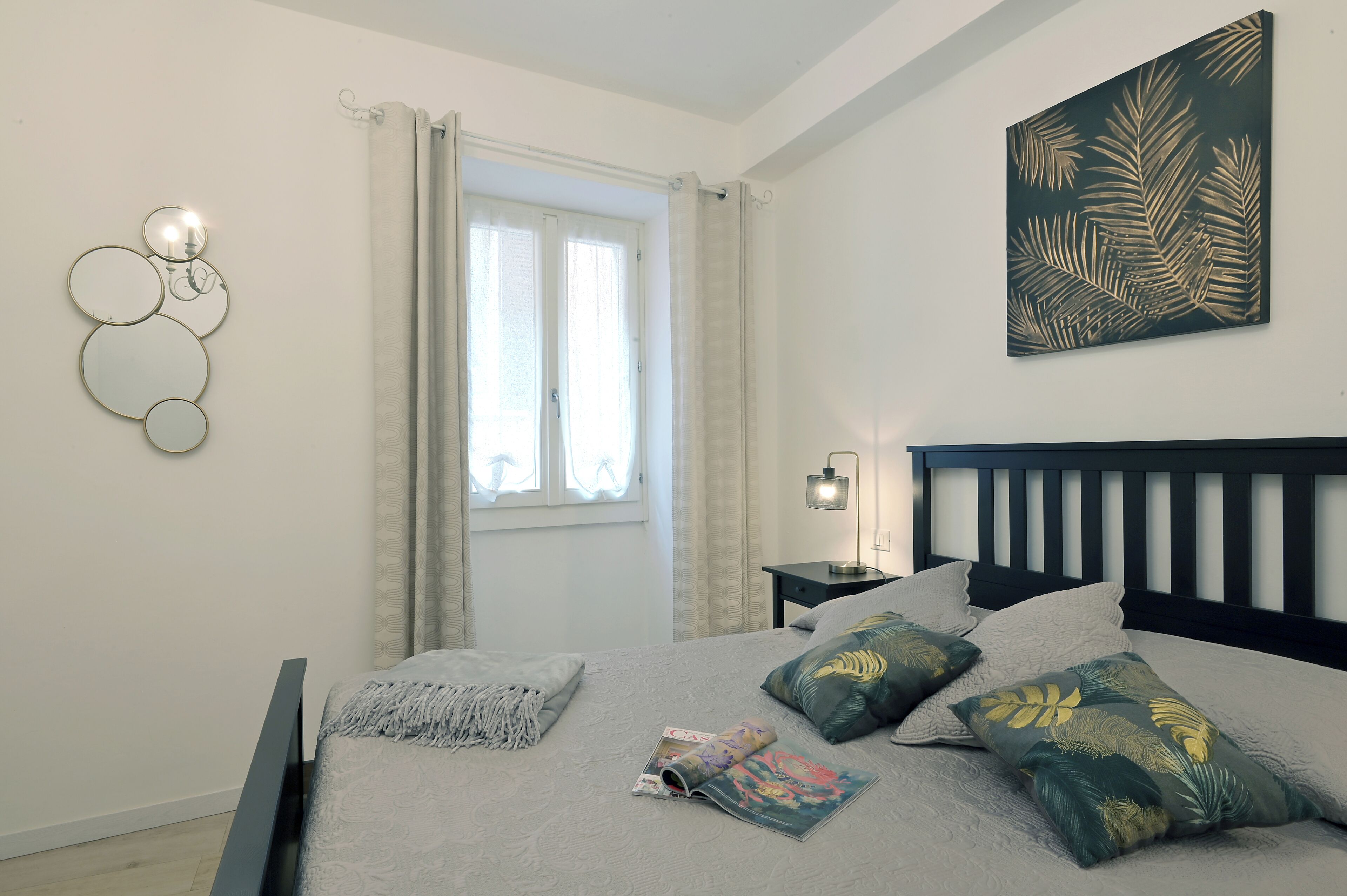 A bedroom with a bed, a nightstand, a wall-mounted lamp, a window with curtains, and a wall art piece.