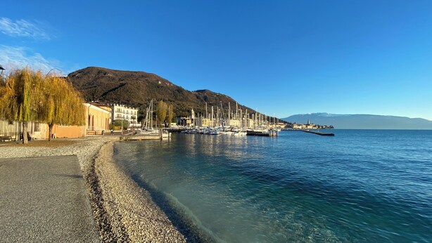 A coastal marina with boats docked, a pebble beach, and buildings along the shore. - Cavour 1 - New apartment at the lake (Salò)