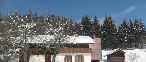 A wooden cabin with a snow-covered roof, surrounded by a wooden fence and snow-covered trees.