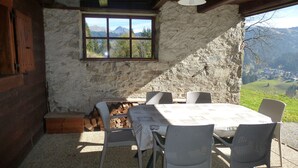 Una cabaña rústica de piedra con interior de madera, una mesa con sillas y vistas a montañas y un pueblo a través de una ventana.