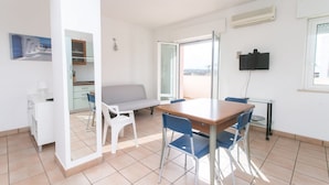 Une cuisine moderne avec des meubles blancs, une table à manger avec des chaises bleues, un canapé et un téléviseur fixé au mur.
