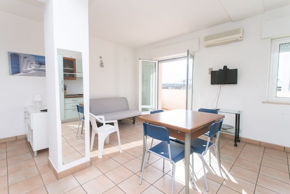 A modern kitchen with white cabinets, a dining table with blue chairs, a sofa, and a TV mounted on the wall.