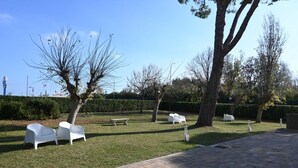 Un espace extérieur aménagé avec une surface pavée, des chaises et des bancs blancs, entouré d’arbres et d’une haie.