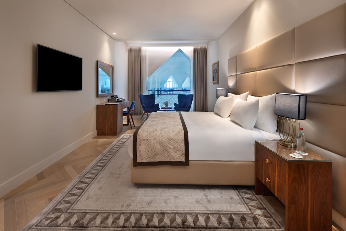 A modern hotel room with a large bed, a TV, a desk with a chair, and a view of a building through the window.