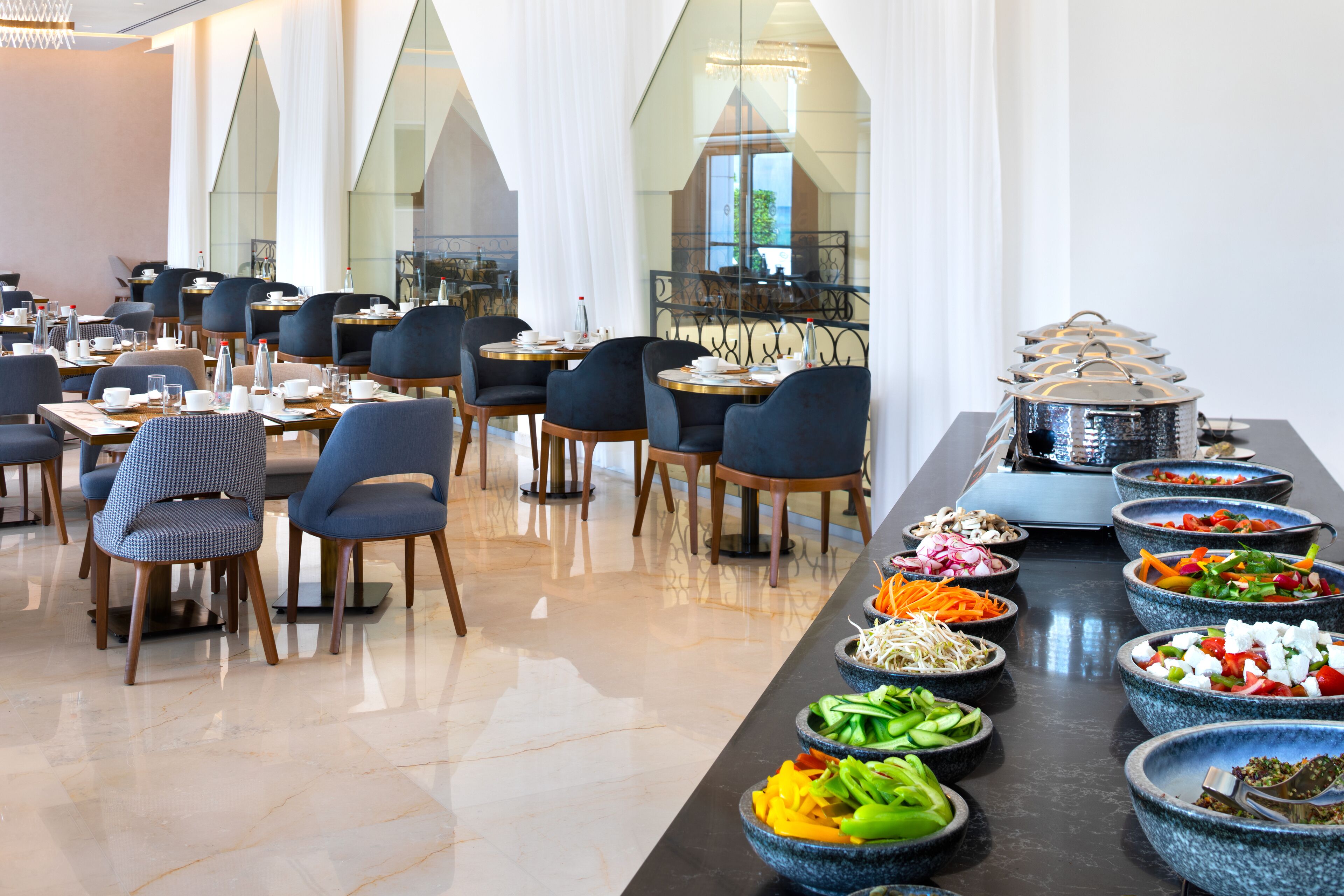 A modern dining area with multiple tables and chairs, a buffet with various dishes, and a view of the outdoors through large windows.