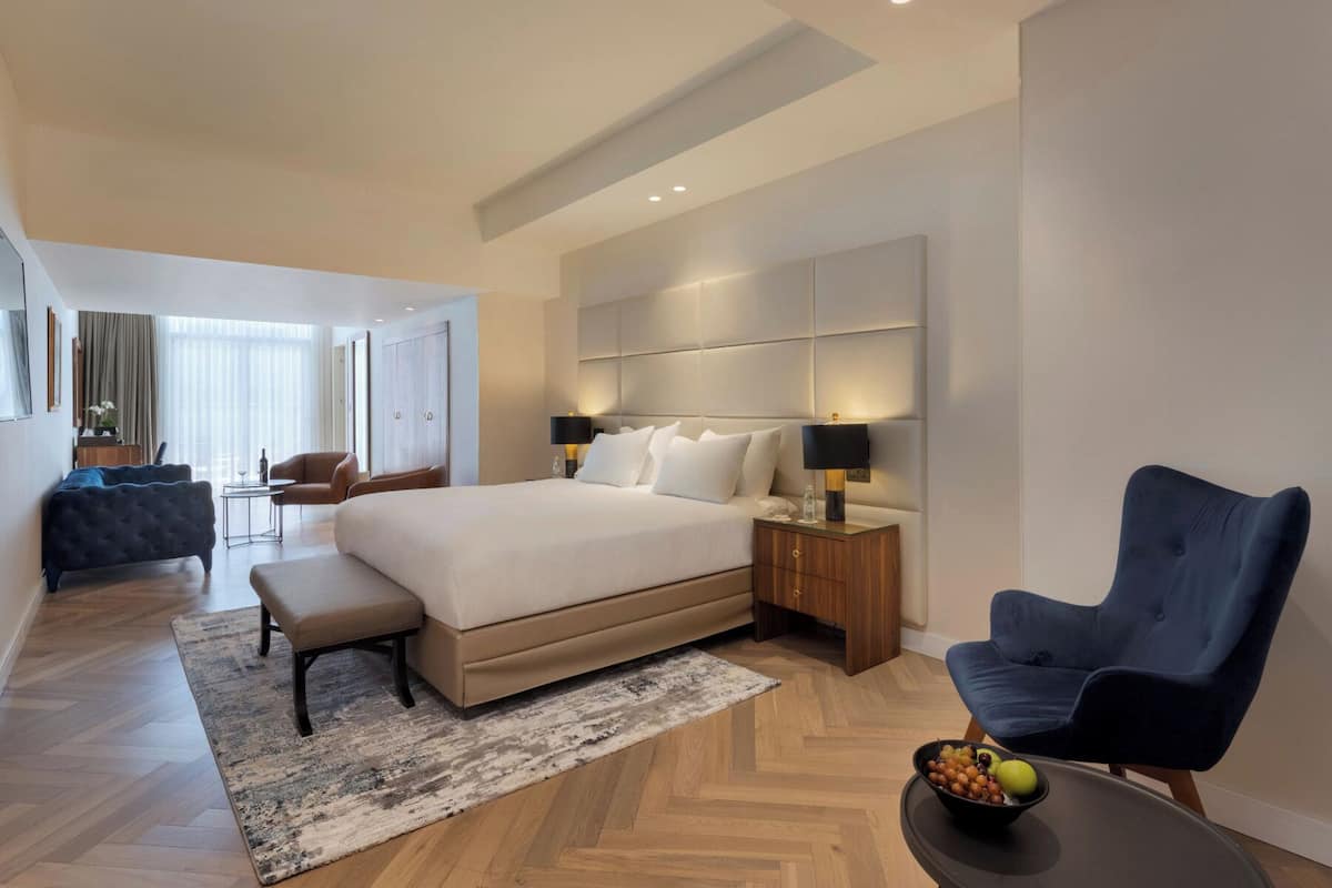 A modern hotel room with a large bed, a desk, a chair, and a dining table with fruit.