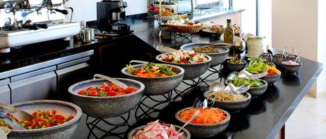 Een buffetopstelling met diverse gerechten in stenen schalen, een koffieapparaat en uitzicht op de stad door grote ramen.