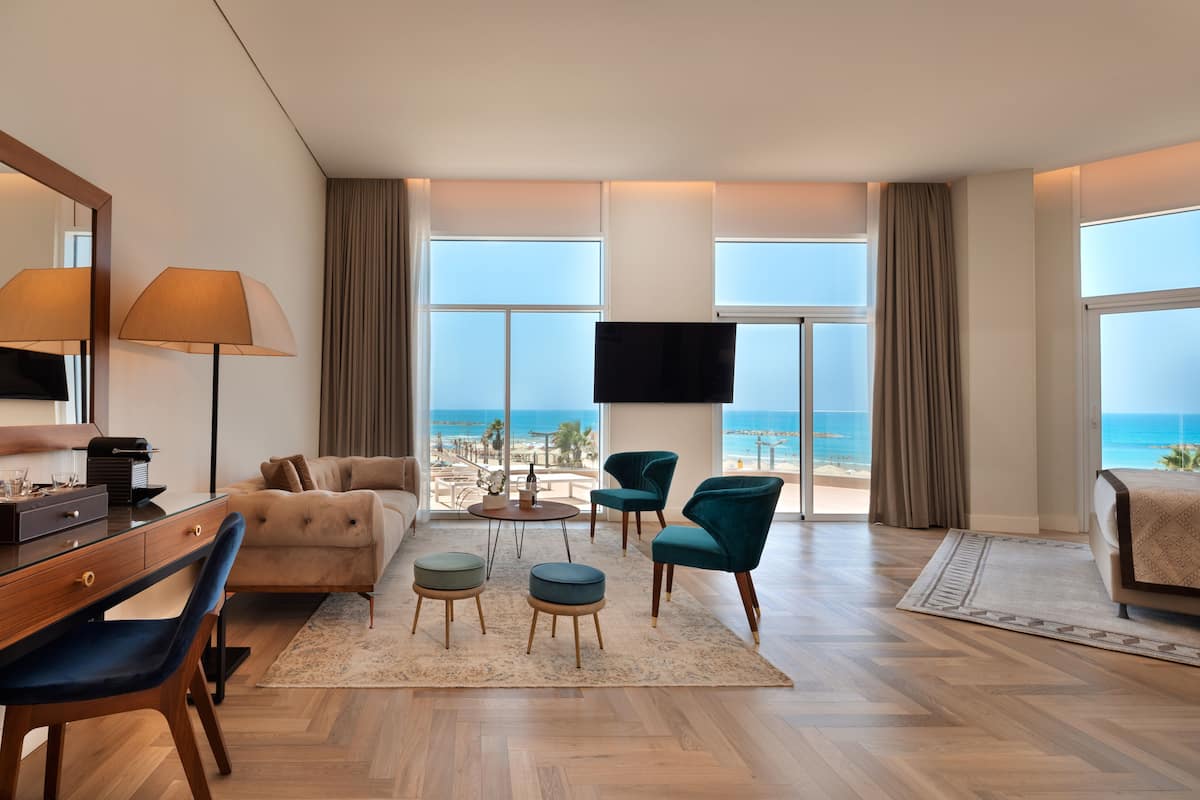 A modern hotel room with a large window offering a beach view, a sofa, a desk with a lamp, and two chairs.